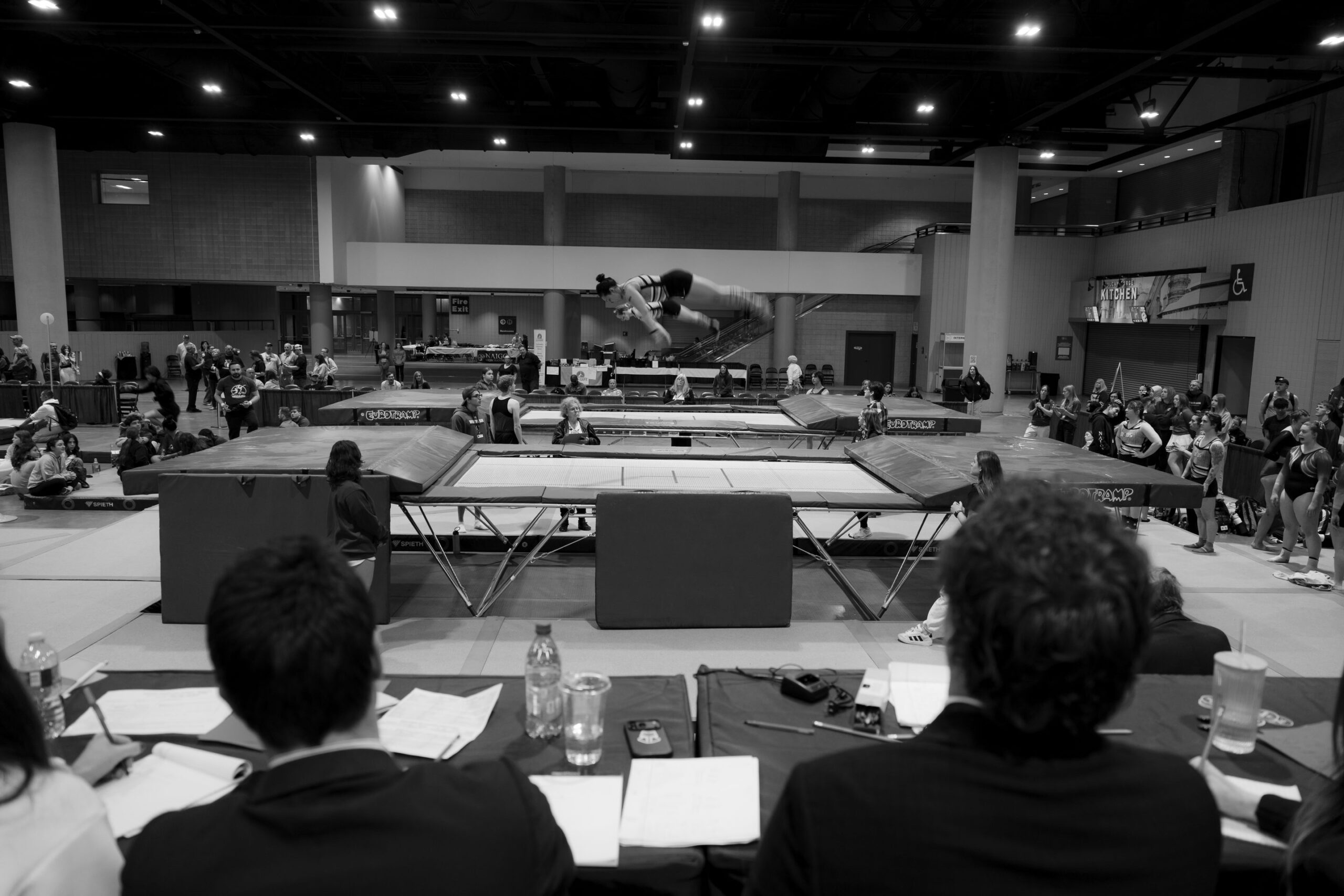 A gymnast in flight above the trampoline as judges watch from the table at UCG Nationals